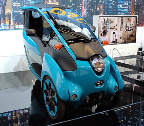 Front of a blue and black narrow three-wheeler with a round headlamp, raised on a display with a city backdrop. Opens a larger view