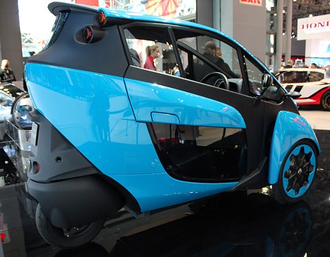 Rear of a blue and black narrow three-wheeler with its single rear wheel, on a show floor. Opens a larger view
