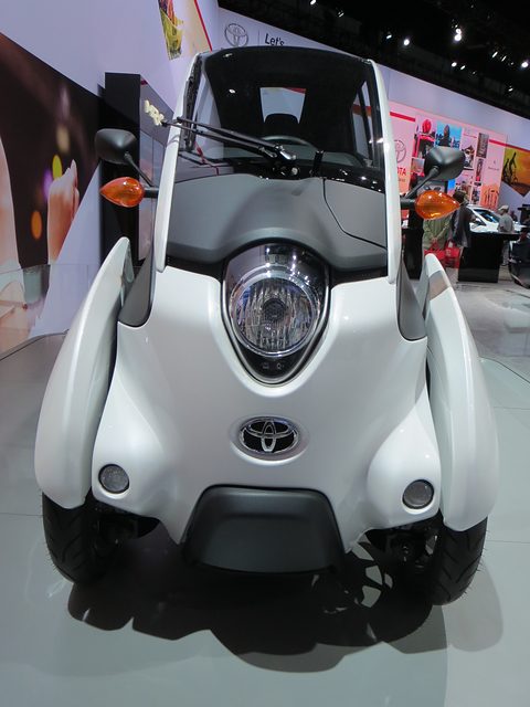 Front of a white and grey narrow three-wheeler with a round headlamp and orange indicators on a show floor. Opens a larger view