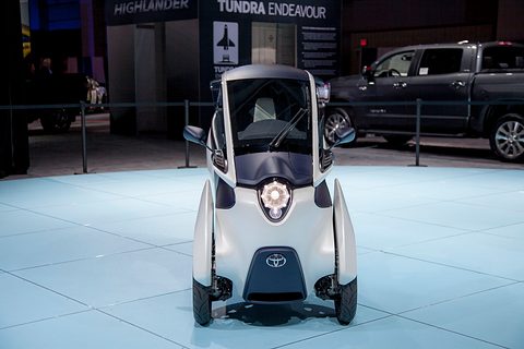 Front of a white narrow three-wheeler with a round headlamp on a pale blue floor, a pick-up truck behind. Opens a larger view