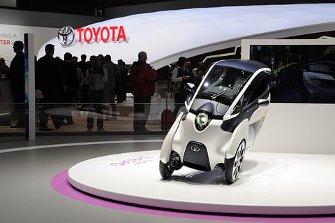 Front of a very narrow white three-wheeler with a round lamp on a turntable before a Toyota sign and a crowd. Opens a larger view