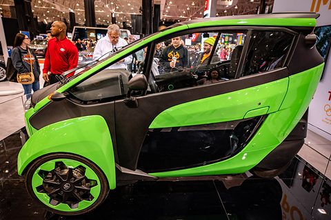 Side view of a lime-green and black narrow three-wheeler on a glossy floor, visitors standing behind it. Opens a larger view