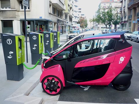 Pink and black narrow three-wheeler connected by a green cable to a row of numbered charging posts in a street.