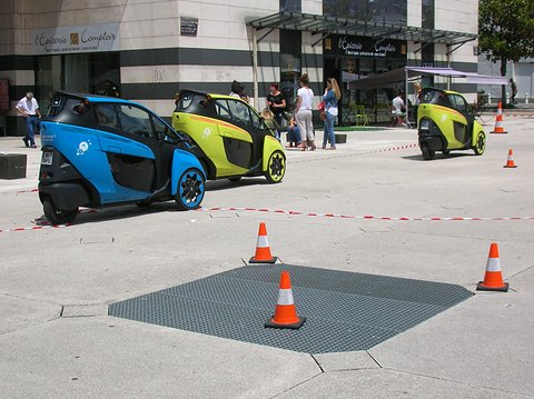 Blue and green narrow three-wheelers on a square marked out with traffic cones, people waiting behind.