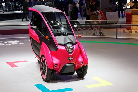 Front of a pink and black narrow three-wheeler with a round chrome headlamp, on a white floor. Opens a larger view