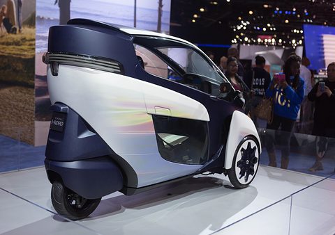 Rear of a white and navy narrow three-wheeler on a glass stand, visitors photographing behind. Opens a larger view