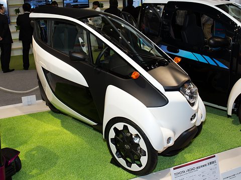 White and black narrow three-wheeler on a green turf stand with a Japanese information sign. Opens a larger view