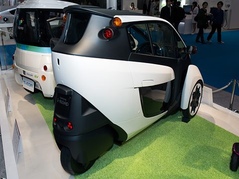 Rear of a white and black narrow three-wheeler on green turf, a Toyota bus behind. Opens a larger view