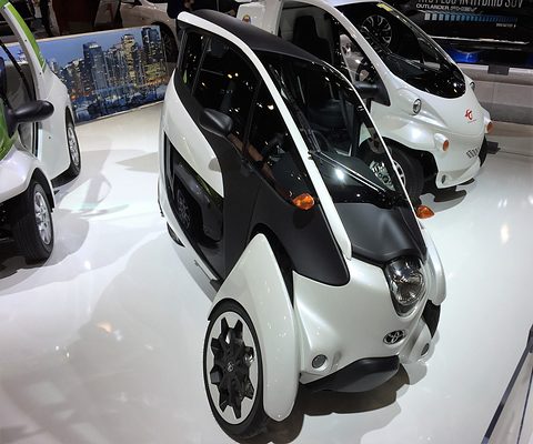 White and black narrow three-wheeler with a single headlamp, seen from low down on a glossy show floor beside another i-Road. Opens a larger view