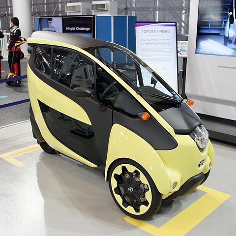Yellow and black narrow three-wheeler on a white floor marked with a yellow box, exhibition panels behind. Opens a larger view