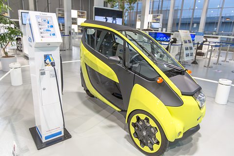 Yellow and black narrow three-wheeler next to a white charging post in a bright showroom.