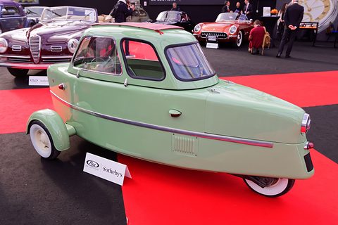 Pale green three-wheeler side-on, chrome strake along its flank and a long pointed tail, on red and black carpet by an RM Sotheby’s card.