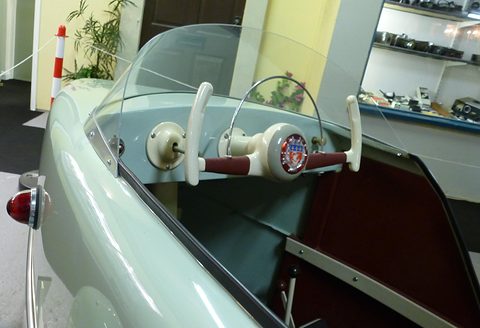 Cream upright handlebar grips on a maroon bar with a central badge, behind a curved windscreen, in a pale turquoise body.