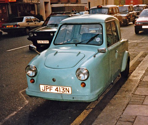 Pale blue single-seat three-wheeler with a hard top parked at a kerb at dusk, cars behind.