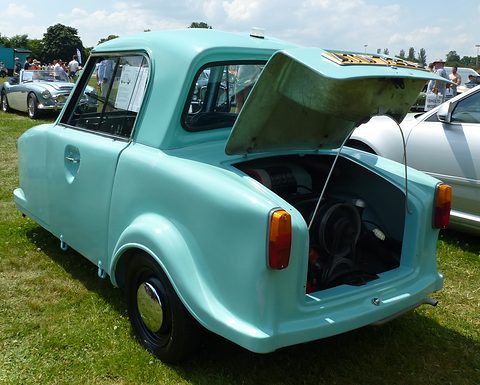 Turquoise single-seat three-wheeler seen from the rear with its tail lid up, on a show field. Opens a larger view