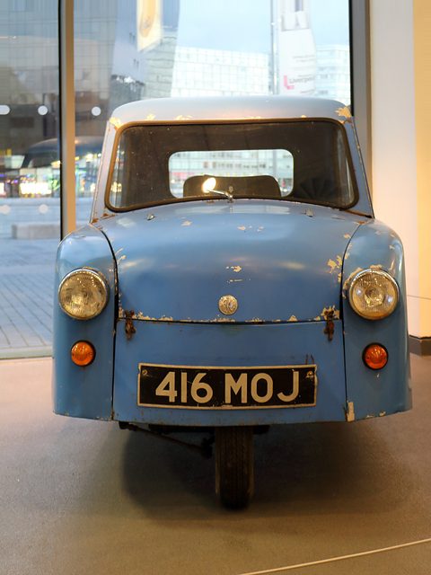 Front view of a worn blue three-wheeler with two headlamps and a narrow single front wheel. Opens a larger view