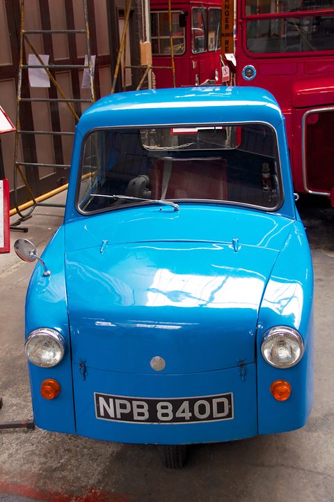 Bright blue single-seat three-wheeler seen head-on beside a red bus in a museum shed. Opens a larger view