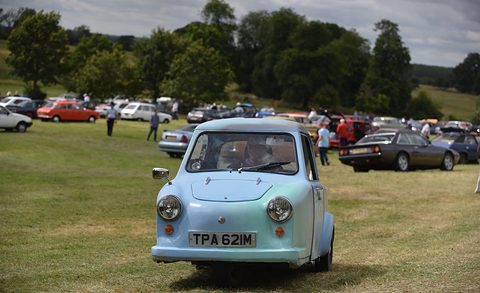 Pale blue single-seat three-wheeler facing the camera on grass, parked cars behind. Opens a larger view