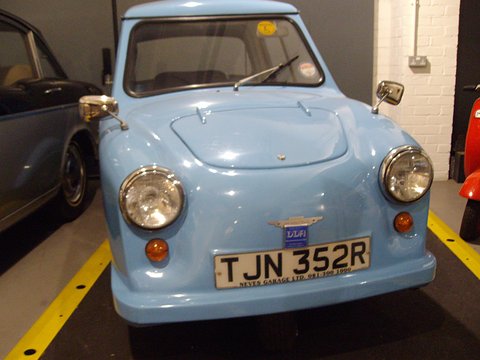 Front view of a pale blue three-wheeler with round headlamps and dealer plate surround. Opens a larger view