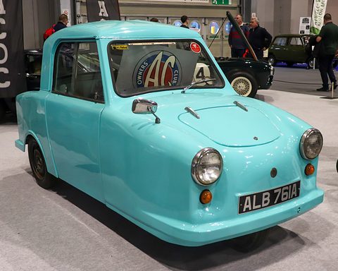 Small turquoise single-seat three-wheeler with a hard top and round headlamps on a grey show floor. Opens a larger view