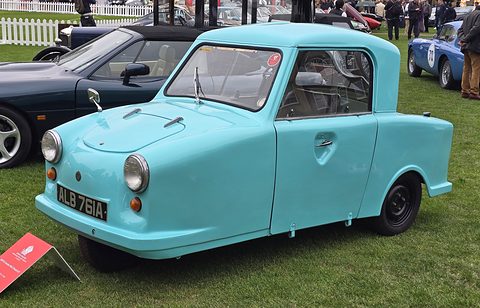 Turquoise single-seat three-wheeler parked on a lawn beside sports cars. Opens a larger view