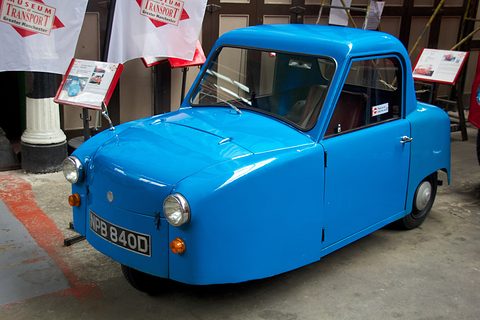 Bright blue single-seat three-wheeler with round headlamps in a museum shed, banners behind. Opens a larger view