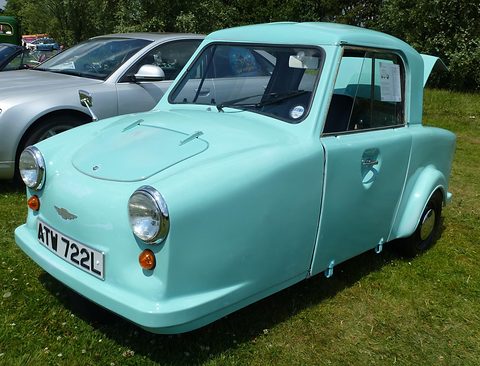 Turquoise single-seat three-wheeler with a hard top parked on grass beside a silver car. Opens a larger view