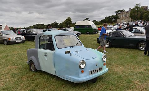 Pale blue single-seat three-wheeler with a grey door on a show field, visitors and cars behind. Opens a larger view