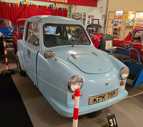 Pale blue single-seat three-wheeler behind a rope and red-and-white posts in a museum. Opens a larger view