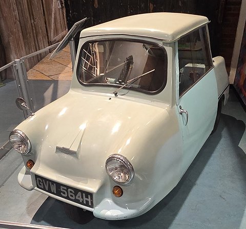 Cream single-seat three-wheeler with its door slid back, in a museum street scene.