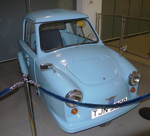 Pale blue single-seat three-wheeler behind a blue rope in a museum, front quarter view. Opens a larger view