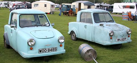Two pale blue single-seat three-wheelers on grass with a milk churn, caravans behind.