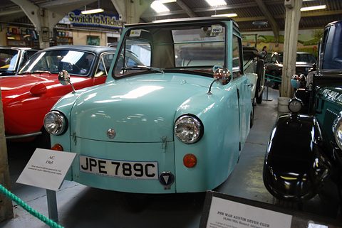 Pale green single-seat three-wheeler with an information card, among cars in a museum shed. Opens a larger view