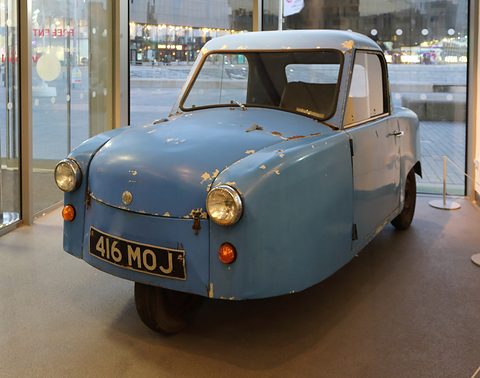 Worn pale blue single-seat three-wheeler with chipped paint in a glass-walled foyer. Opens a larger view