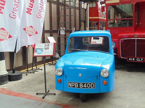 Bright blue three-wheeler with an information stand, flags and a red double-deck bus behind. Opens a larger view