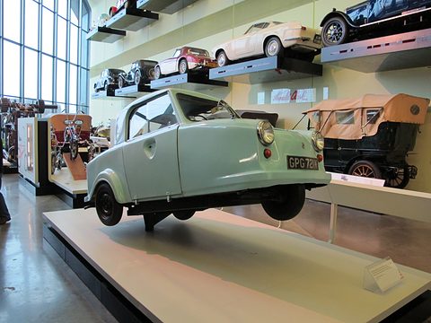Pale green three-wheeler on a white plinth, cars mounted on shelves on the wall above. Opens a larger view