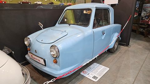 Pale blue single-seat three-wheeler behind a red and white chain in a museum hall. Opens a larger view