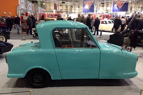 Side view of a turquoise single-seat three-wheeler with a short hard top in a busy show hall. Opens a larger view