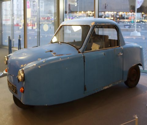 Side view of a chipped blue single-seat three-wheeler with a hard top, city buildings beyond the glass. Opens a larger view