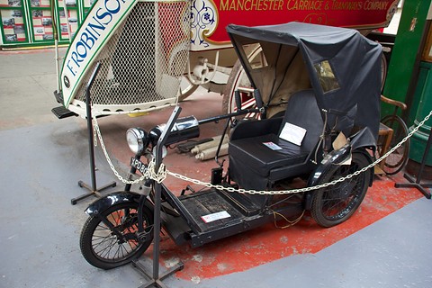 Side view of a black open invalid tricycle with a hood and spoked wheels behind a chain. Opens a larger view