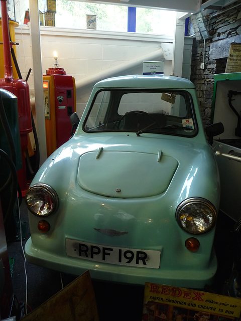 Pale green single-seat three-wheeler seen head-on among petrol pumps and museum displays. Opens a larger view