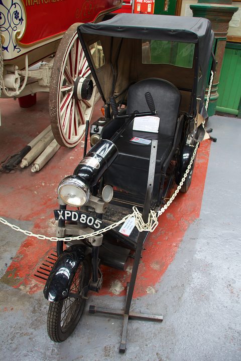 Black open tricycle with a padded seat, folding hood and front lamp behind a chain.