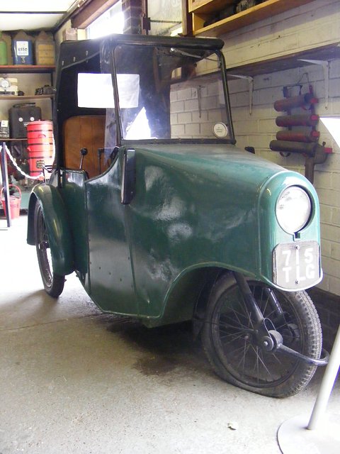 Dark green three-wheeled invalid carriage with a black fabric hood and spoked wheels in a workshop.