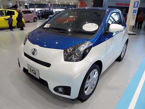 White city car with a blue roof and bonnet on a showroom floor, other cars and visitors behind. Opens a larger view