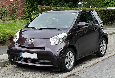 Dark aubergine city car with silver wheels parked on a pavement in front of a garden hedge, plate blanked. Opens a larger view