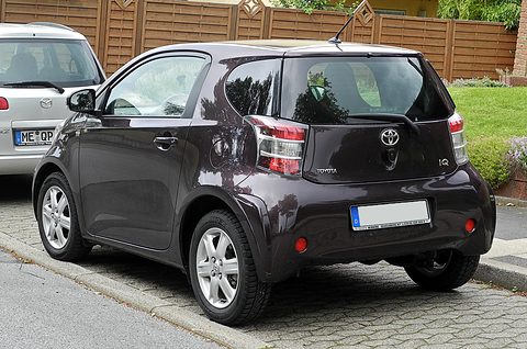Rear of a dark aubergine city car with tall tail lamps parked on block paving by a wooden fence. Opens a larger view