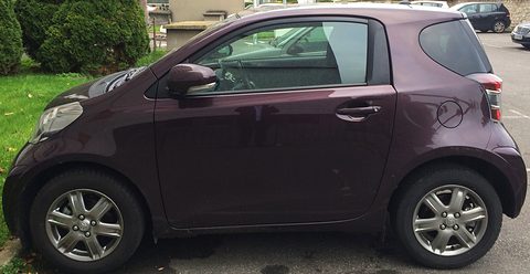Side view of a dark purple three-door city car with a very short wheelbase and silver wheels, parked by bushes. Opens a larger view