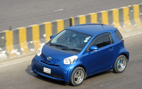 Blue city car driving fast past a black and yellow striped road divider. Opens a larger view