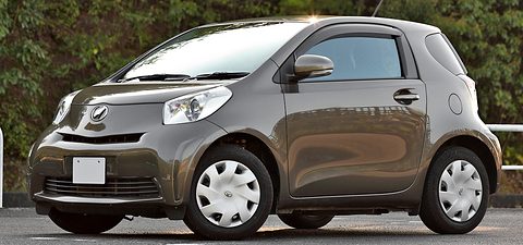 Bronze-brown short city car with plastic wheel covers parked in front of a wooded hillside, plate blanked. Opens a larger view