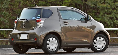 Rear of a bronze-brown short city car with stickers in its rear window, parked by a railing and trees. Opens a larger view
